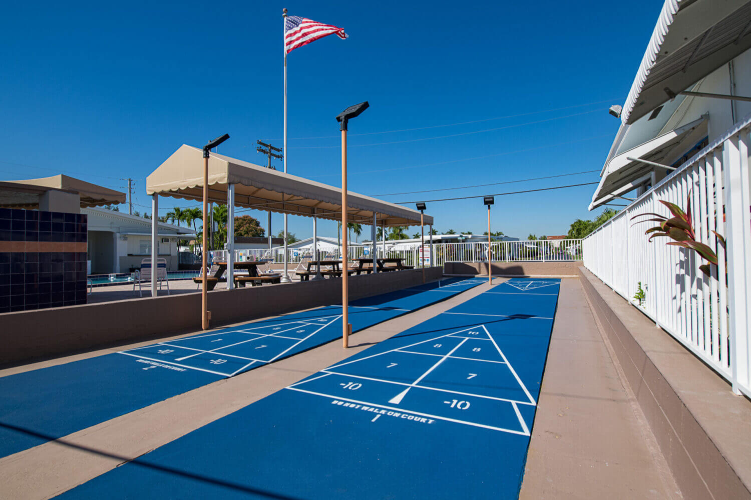 Blue outfdoor shuffleboard courts with pole lamps at a fenced area.