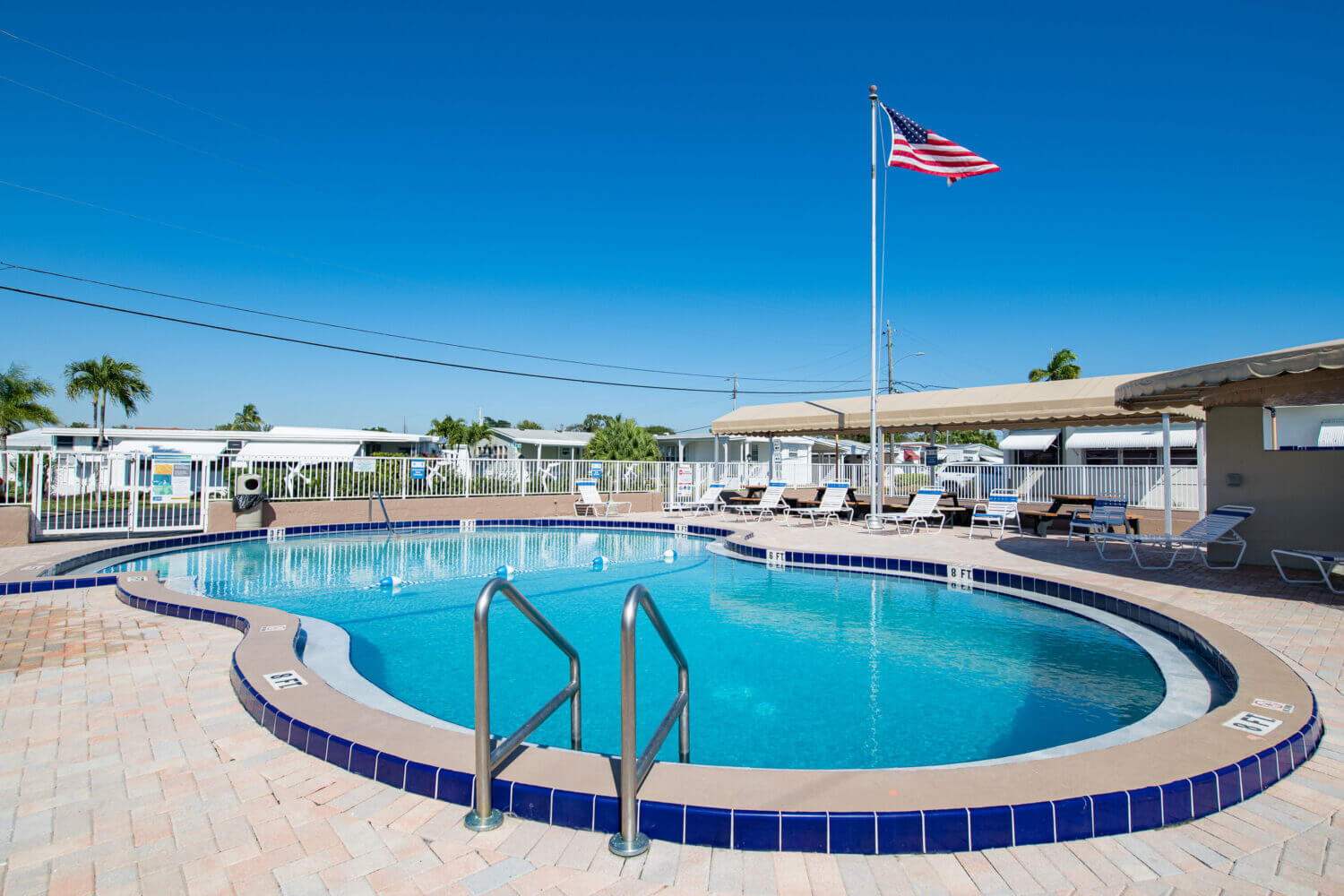 Swimming pool in a fenced area with sunbeds under clear skies and the American flag on a pole in the background.